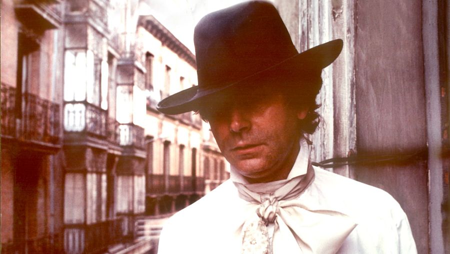 Imagen promocional de "El loco. Los silencios de Quintero - Episodio 2".  Muestra al actor español Imanol Arias con sombrero y pañuelo al cuello, en una calle con edificios antiguos.