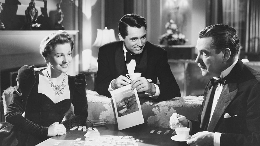 Cary Grant, Joan Fontaine y Nigel Bruce en una escena de "Sospecha", sentados a una mesa, con tazas y una fotografía.