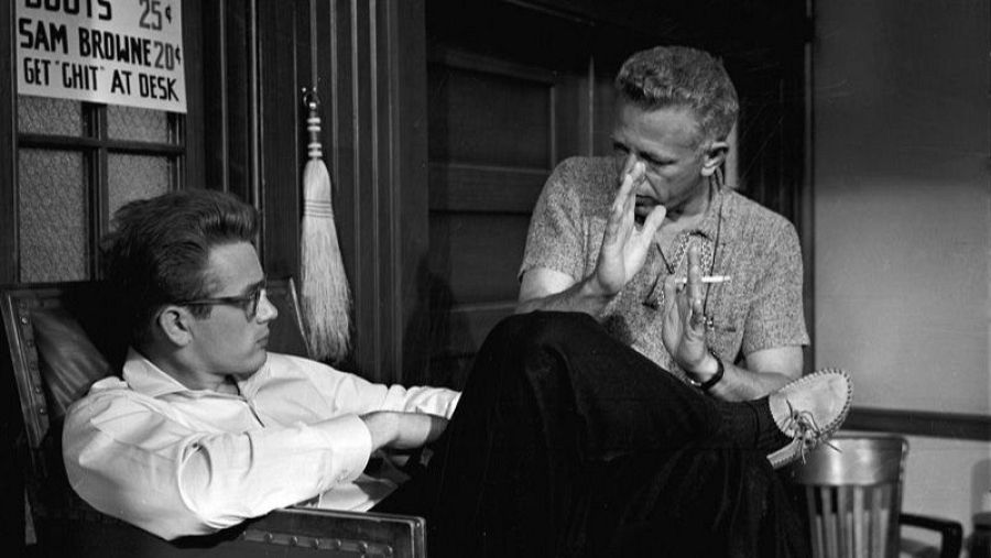 James Dean y Nicholas Ray durante el rodaje de "Rebelde sin causa". Dean, con camisa blanca y gafas, está sentado; Ray gesticula mientras fuma.