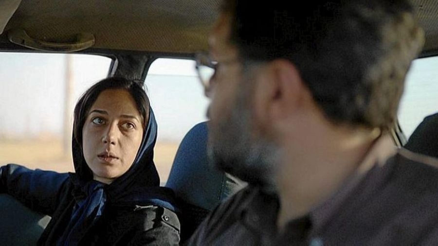 Mujer con pañuelo y hombre con gafas en un coche. Paisaje desértico visible por la ventana. Escena de la película "Araña Sagrada".