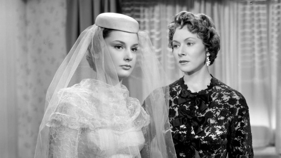 Dos mujeres con vestidos elegantes en una escena en blanco y negro de la película "Alta Costura". Una viste blanco con velo, la otra un vestido oscuro de encaje.