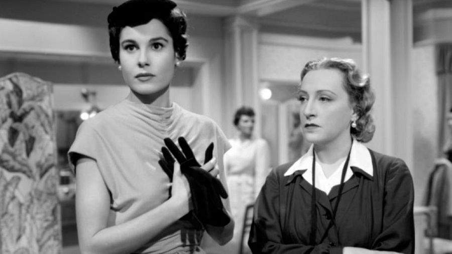 Dos mujeres, una con vestido y guantes negros, la otra con camisa blanca y chaqueta oscura, en una escena de la película "Alta Costura".