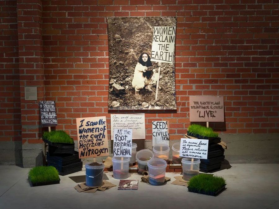 Instalación "SPROUTIME IS NOW!" de Leslie Labowitz Starus (1980-2023): fotografía de mujer con cartel "WOMEN RECLAIM THE EARTH", carteles con lemas ecofeministas, plantas y tierra. Ecofeminismo y resistencia ambiental.