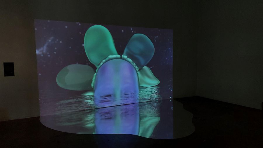 Proyección de "Deep Time Dance" (2024) de Kira Xonorika: figura con cabeza sumergida en líquido reflectante, fondo estrellado. Efecto espejo en el suelo.