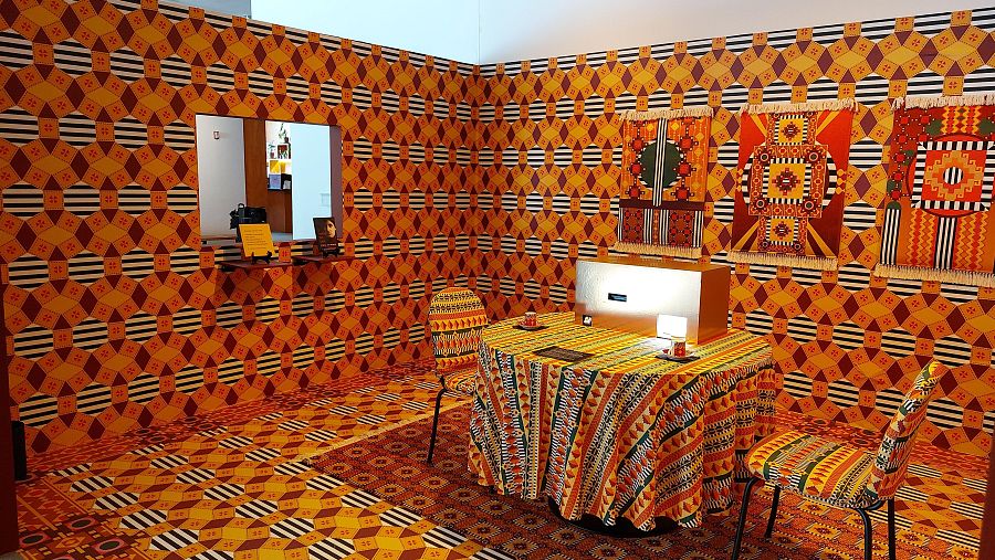 Instalación "One Who Looks at the Cup": habitación con patrones geométricos vibrantes en naranja, amarillo, marrón y negro. Mesa, sillas y tapices con diseños intrincados.