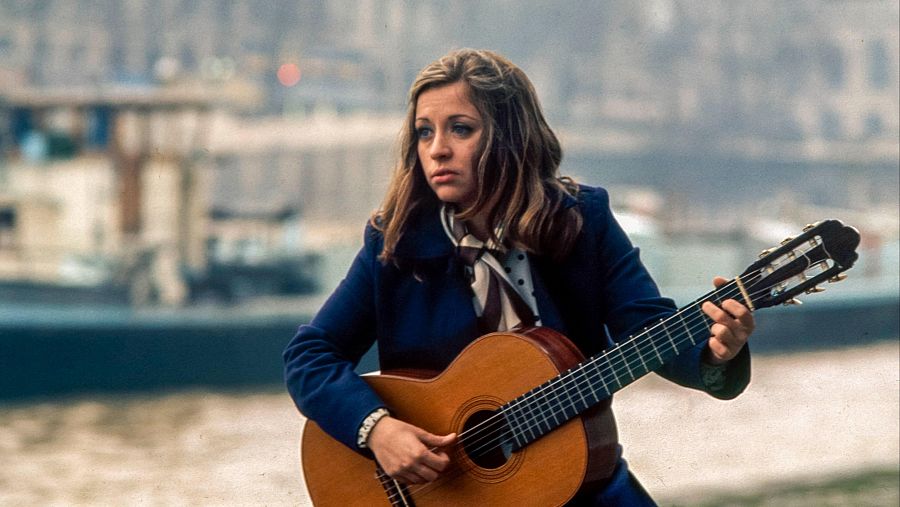 Mari Trini tocando la guitarra en París, junto a un río. Lleva una chaqueta azul y pañuelo estampado.