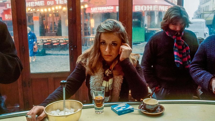 Mari Trini en un café parisino.  Sentada en la barra, disfruta de un café con compañía.  Se ven bebidas, azúcar y tabaco en la barra.