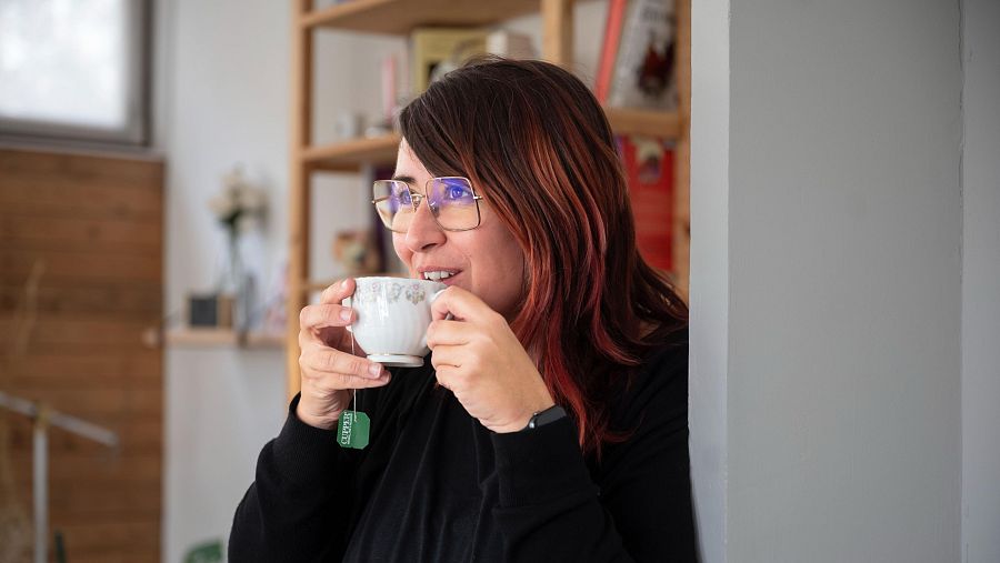 Elena Crespi bebe té, sonriendo. Lleva gafas y una blusa negra.  Fondo con libros y texto: "Elena Crespi vincula el feminisme amb la revolució sexual"