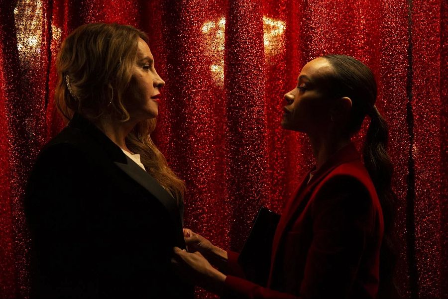 Karla Sofía Gascón y Zoe Saldaña en una escena de "Emilia Pérez".  Ambas de perfil, con un telón rojo detrás. Zoe ajusta la chaqueta de Karla.