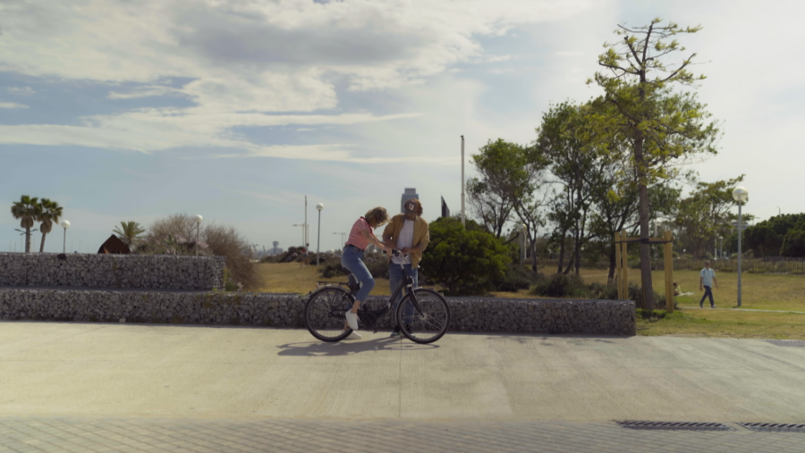 Santi Millán, con camisa mostaza, ayuda a una mujer en bici en un parque. Día soleado, fondo con árboles y costa.