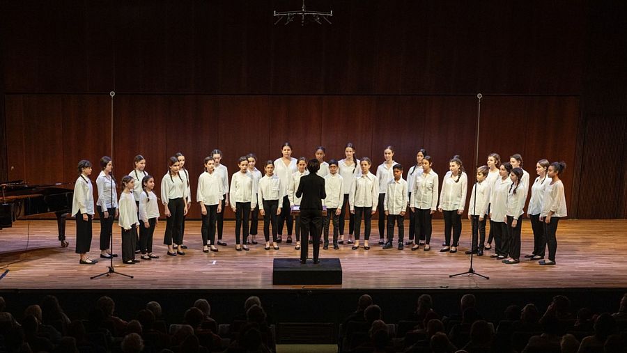 Actuación de un coro infantil uniformado en un escenario con piano y micrófonos. El director, de espaldas, guía a los pequeños cantantes.