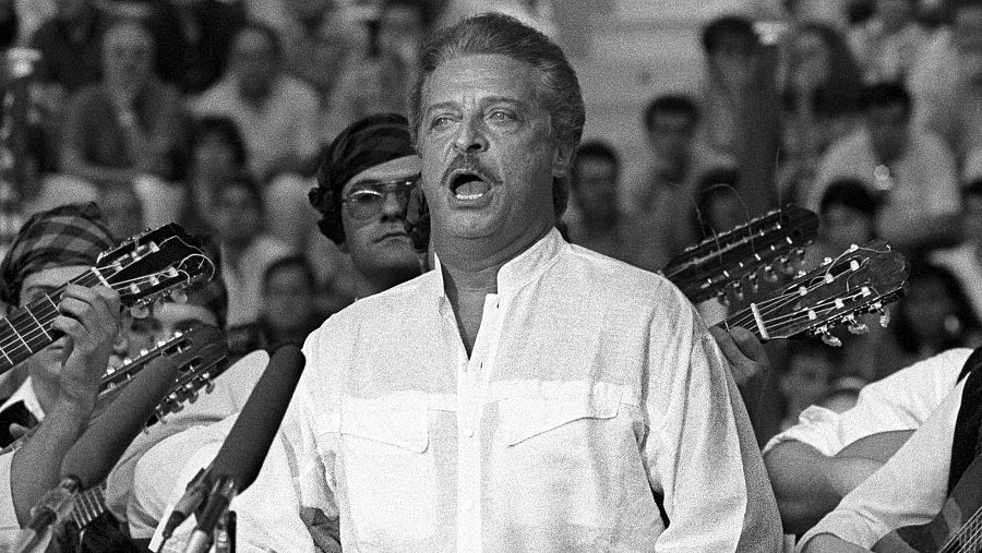 Alfredo Kraus, en blanco y negro, cantando al micrófono; viste camisa blanca.  Músicos con guitarras al fondo.