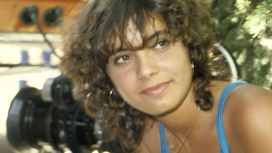 Retrato de Sonia Martínez con cabello rizado castaño y camiseta azul, tomada al aire libre cerca de una cámara.  Posiblemente de un documental.