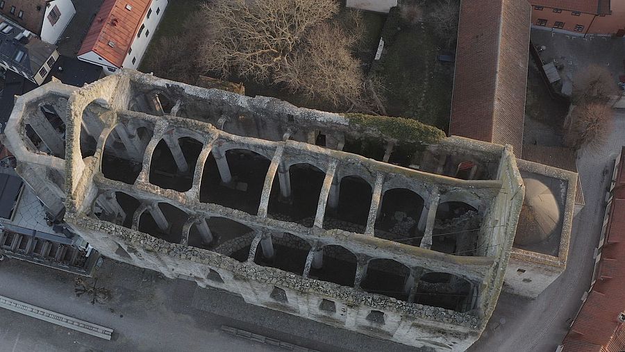 Vista aérea de ruinas de iglesia con arcos de piedra, paredes derruidas y una sección con techo conservado.  Se aprecian edificios adyacentes y un árbol sin hojas.