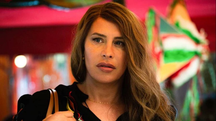 Karla Sofía Gascón en escena de "Emilia Pérez", con blusa oscura, bolso beige y pelo castaño ondulado. Fondo festivo con colores verde, blanco y rojo.
