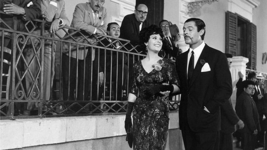 Marcello Mastroianni y Stefania Sandrelli, elegantemente vestidos, son el centro de atención en esta escena de "Divorcio a la italiana".  Un balcón con hombres observando y espectadores completan la imagen.