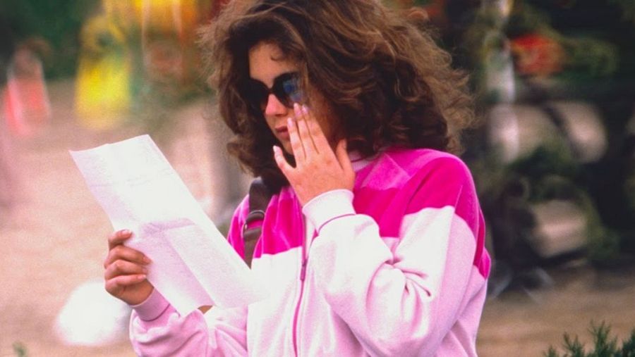 Mujer con chaqueta deportiva rosa y blanca lee un documento, tocándose la cara. Fondo exterior desenfocado. Posible conexión con documentales.