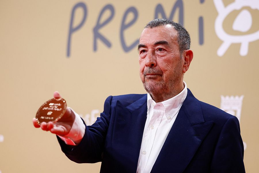 José Luis Garci muestra la Medalla de Oro de los Premios Forqué.  Americana azul marino y camisa blanca; fondo beige claro con parte de la palabra "PREMI" visible.