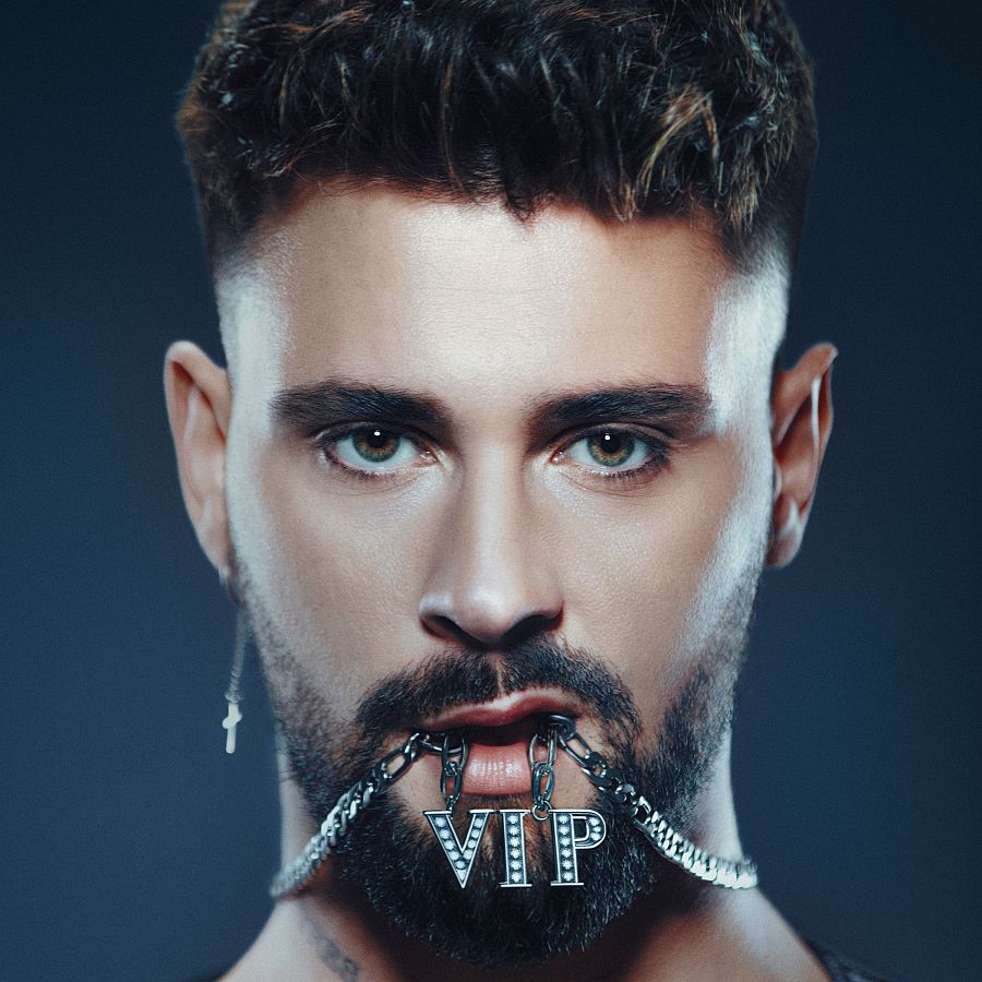 Primer plano de J Kbello sosteniendo una cadena con un colgante "VIP" con brillantes. Pelo corto, oscuro, barba y mirada directa a cámara.