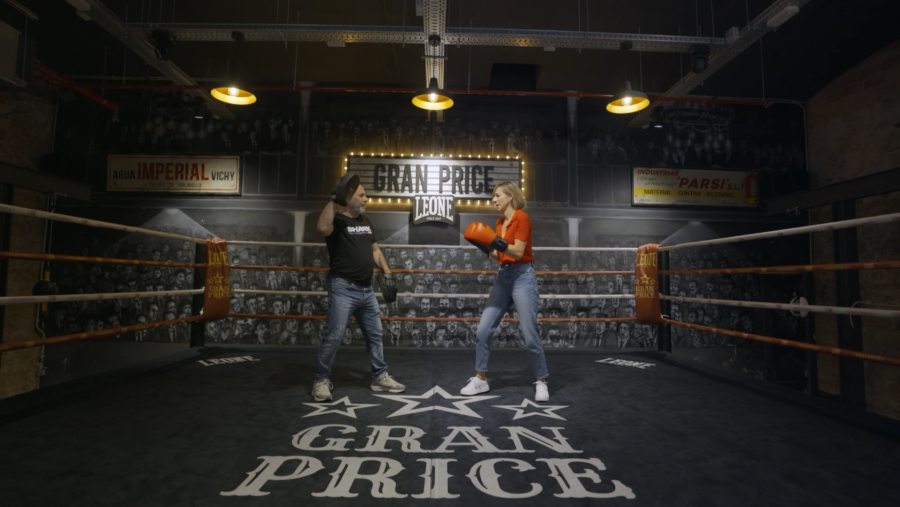 Combate de boxeo entre hombre con camiseta negra y mujer con camiseta roja y guantes rojos.  El ring tiene publicidad antigua y se ve "GRAN PRICE" en el suelo.