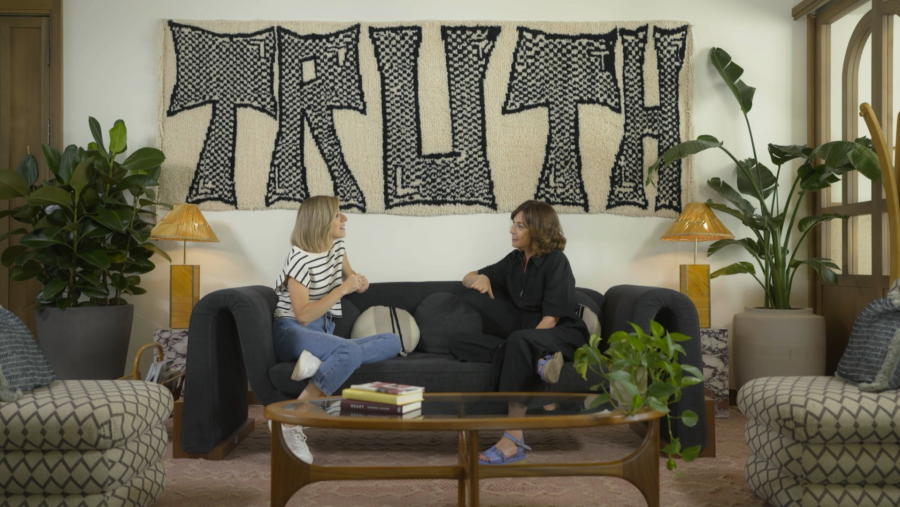 Dos mujeres sentadas en un sofá, con lámparas y plantas alrededor.  Una pieza decorativa con la palabra "TRUTH" en la pared.