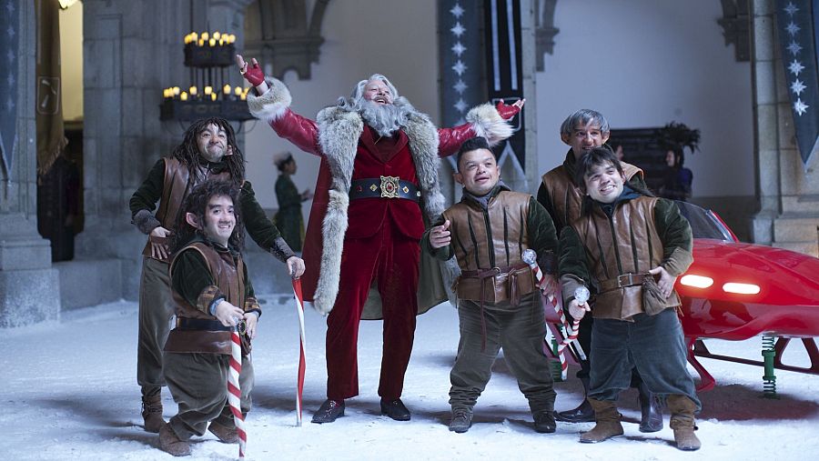 Santa Claus, con traje rojo y barba blanca, rodeado de cuatro personajes medievales con bastones de caramelo, en un patio nevado con un vehículo rojo. Escena de la película "Reyes contra Santa".