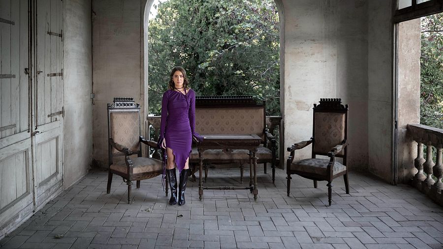 Cantante con vestido morado y botas negras en un porche antiguo con sillas y sofá. Ambiente sombrío y misterioso.