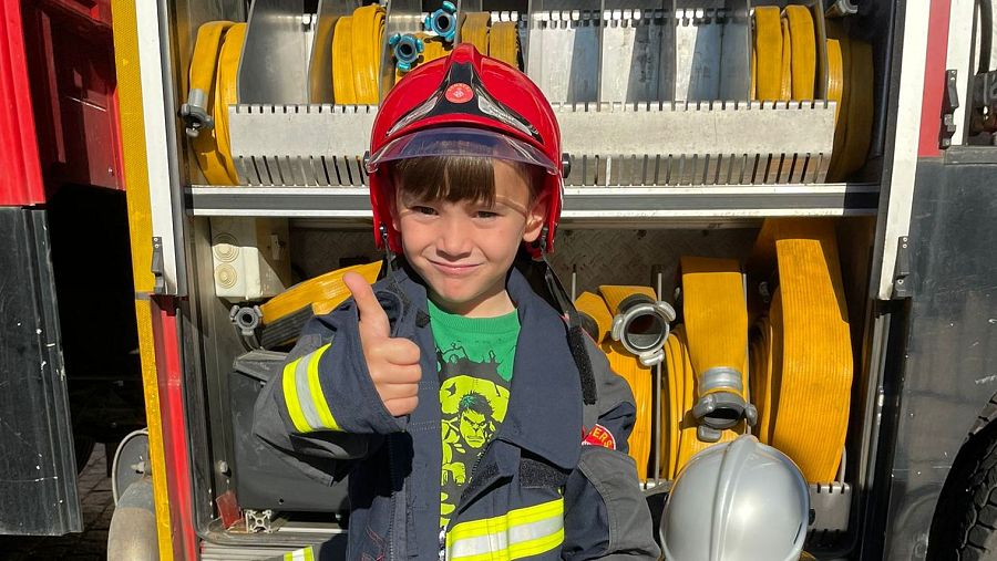 Niño con uniforme de bombero, casco rojo y pulgar arriba, sostiene un casco plateado. Fondo: camión de bomberos con mangueras amarillas.