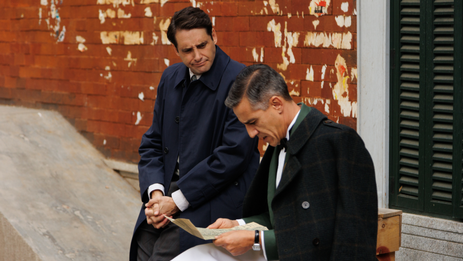 Dos hombres, uno con abrigo oscuro y el otro con abrigo oscuro y corbata, examinan un documento ante una pared de ladrillo.  Escena de época.