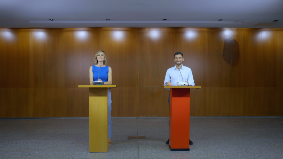 Rueda de prensa con dos personas sonrientes tras atriles con micrófonos, en un escenario iluminado con focos sobre pared de madera.
