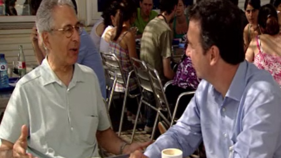 Encuentro informal entre dos hombres en una terraza. Uno lleva camisa clara y gafas; el otro, camisa azul. Conversación animada.