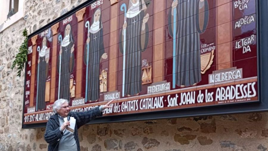 Mural de azulejos con figuras femeninas y sus nombres,  inscripción "Abadesses dels Comtats Catalans - Sant Joan de les Abadesses", y persona explicándolo con micrófono.