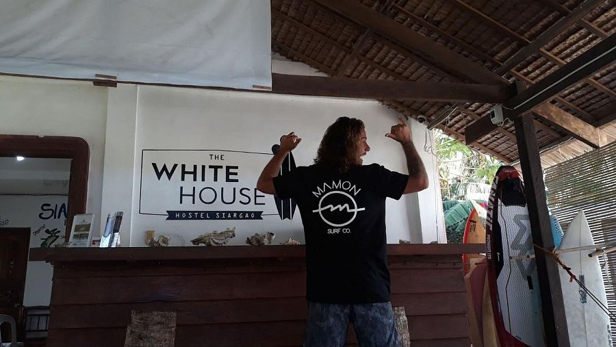 Diego Bello muestra su aprobación con los pulgares arriba en un albergue con tablas de surf. Lleva una camiseta negra con el logo "MAMON SURF CO.".