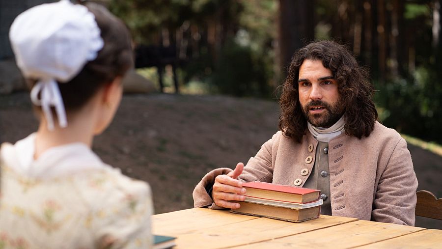 Hombre y mujer, vestidos de época, conversan a una mesa con libros antiguos en un entorno natural.  Posible escena de "Valle Salvaje".