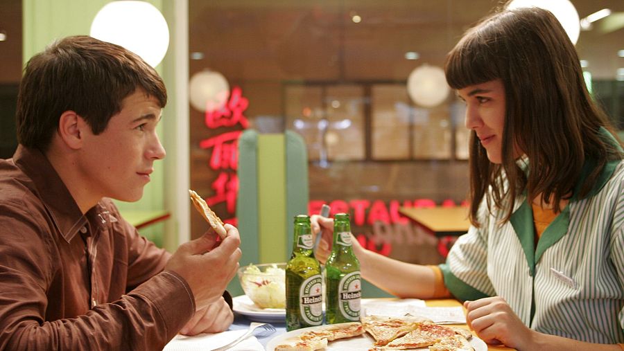 En un restaurante, un hombre con camisa marrón y una mujer con uniforme a rayas comparten pizza y cerveza Heineken.  Escena informal de la película "Cabeza de perro".