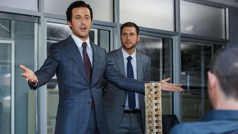 Ryan Gosling, con traje azul oscuro, gesticula enérgicamente en una escena que incluye a otros dos hombres y una torre de bloques de madera.