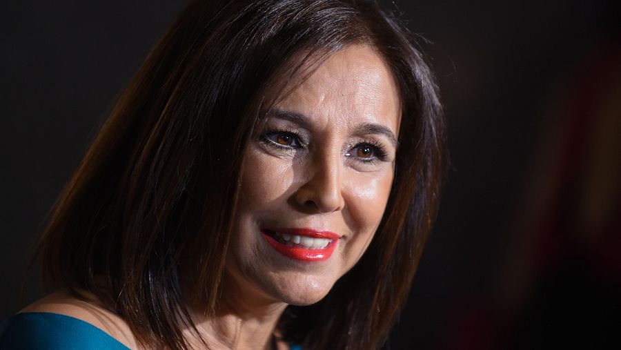 Presentadora con pelo castaño liso hasta los hombros, labios rojos y blusa oscura sonriendo ligeramente sobre fondo oscuro.