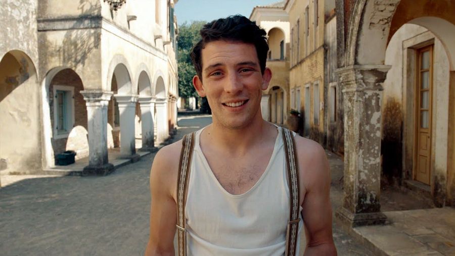 Josh O'Connor, con cabello corto y camiseta blanca, sonríe en una calle estrecha con edificios de estilo mediterráneo.