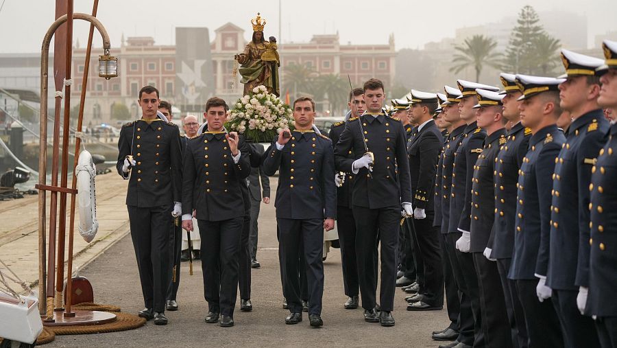 Guardamarinas en uniforme portan una imagen religiosa adornada con flores blancas en una procesión marítima. Se observan elementos náuticos en el fondo.
