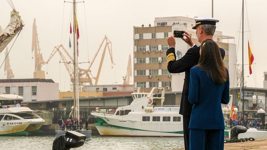 El rey Felipe VI y la princesa Leonor, en trajes azules, en un puerto. El rey fotografía con su móvil, con barcos y edificios al fondo.