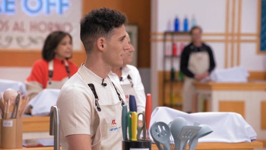 Pol Espargaró, con delantal beige y logo de 'Bake Off', participa en una prueba de cocina.  Utensilios de cocina en la mesa, fondo con otras personas.