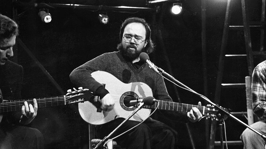 Gato Pérez, con guitarra y micrófono, canta en un escenario oscuro junto a otros dos guitarristas durante una actuación en vivo. Imagen en blanco y negro.