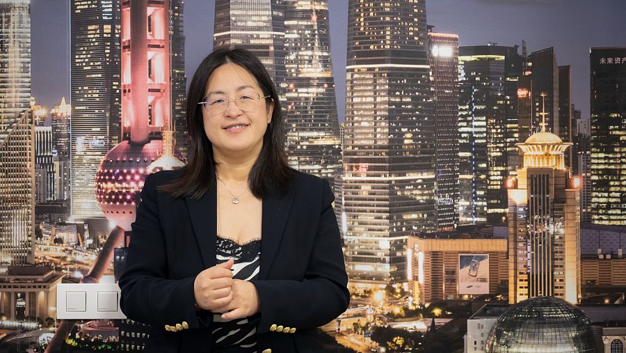 Mujer asiática con gafas y chaqueta negra sobre blusa de cebra, con las manos juntas, frente a un fondo nocturno de rascacielos iluminados.