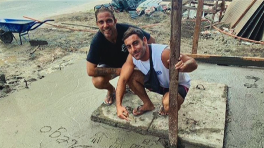 Dos hombres agachados en una obra, uno señala una fecha ("05/24") en cemento fresco. Materiales de construcción visibles.