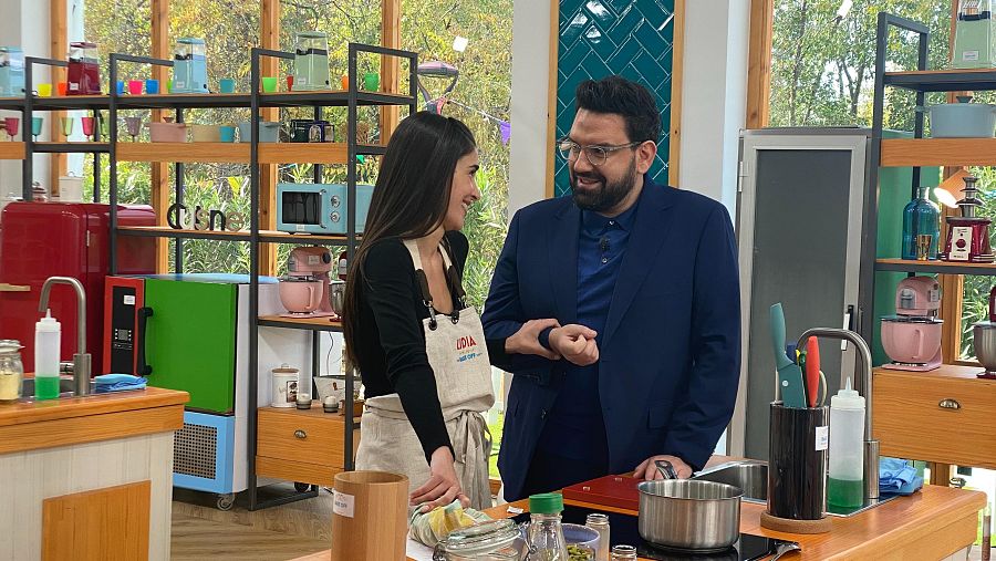 Lidia Torrent, con delantal, y Damián Betular, con americana azul, conversan en una cocina colorida llena de utensilios mientras preparan comida.