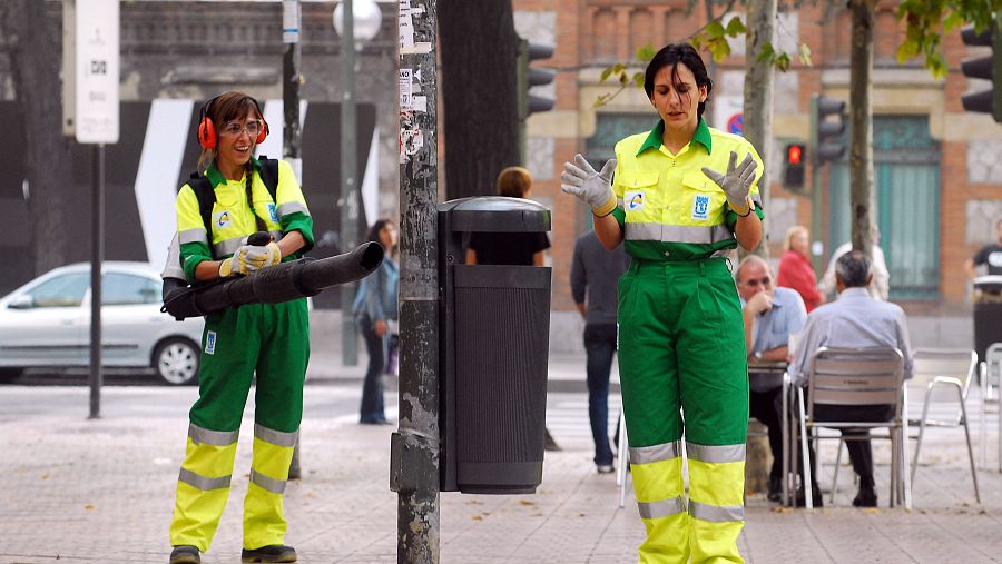 Dos limpiadoras, una con soplador, trabajan en una zona pública con gente al fondo.  En la parte superior: "UNA PALABRA TUYA (7) Somos cine".