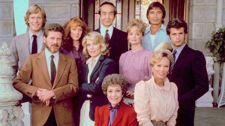 Fotografía del elenco de "Falcon Crest", diez personas vestidas con atuendos de los 80, posando formalmente ante una gran entrada con columnas.