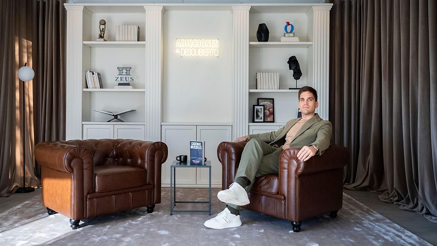 Ex adicto y terapeuta en su consulta, con sillones, estanterías con libros sobre adicciones y un letrero de neón que dice "Adicciones al descubierto".