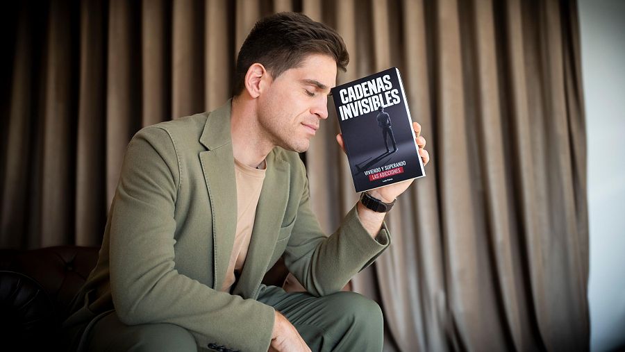 Autor sentado en sillón, traje verde oliva, sosteniendo su libro con portada negra y título "CADENAS INVISIBLES" en letras blancas.  Atmósfera tranquila y reflexiva.