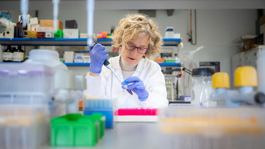 Investigadora en genética manipulando una pipeta y una placa de pocillos en un laboratorio. Lleva bata blanca y guantes azules.